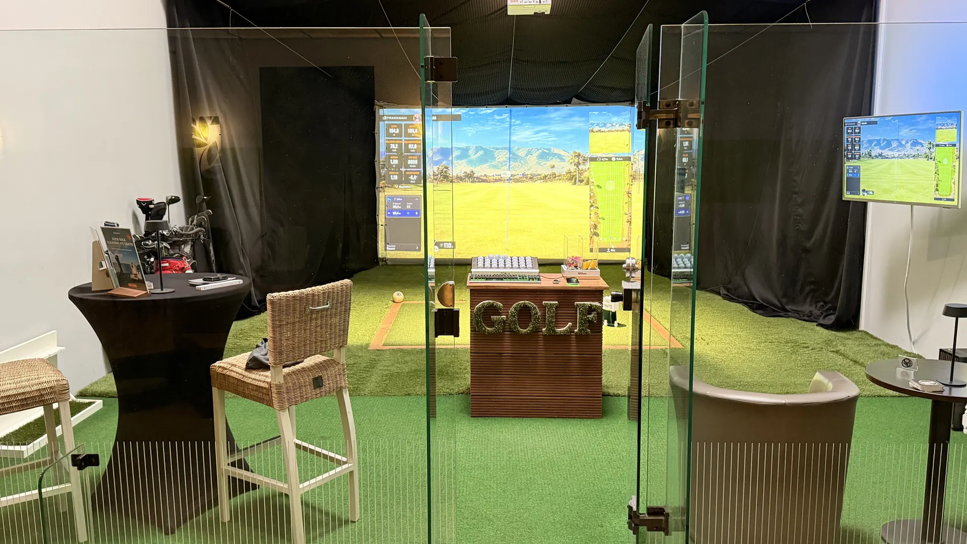 Blick durch die Glastüren in den Green Corner: Abschlagmatte, Trackman-Leinwand mit Golfplatz und Monitor zur Schlaganalyse