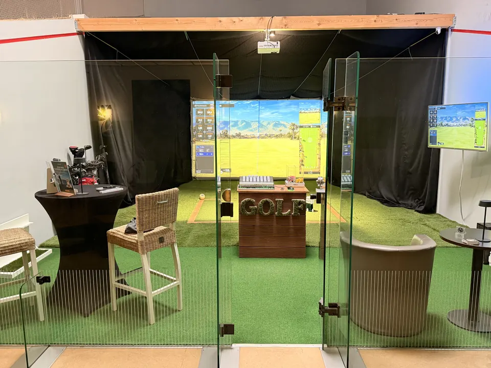 Blick durch die Glastüren in den Green Corner mit Leinwand, Abschlagmatte und Monitor