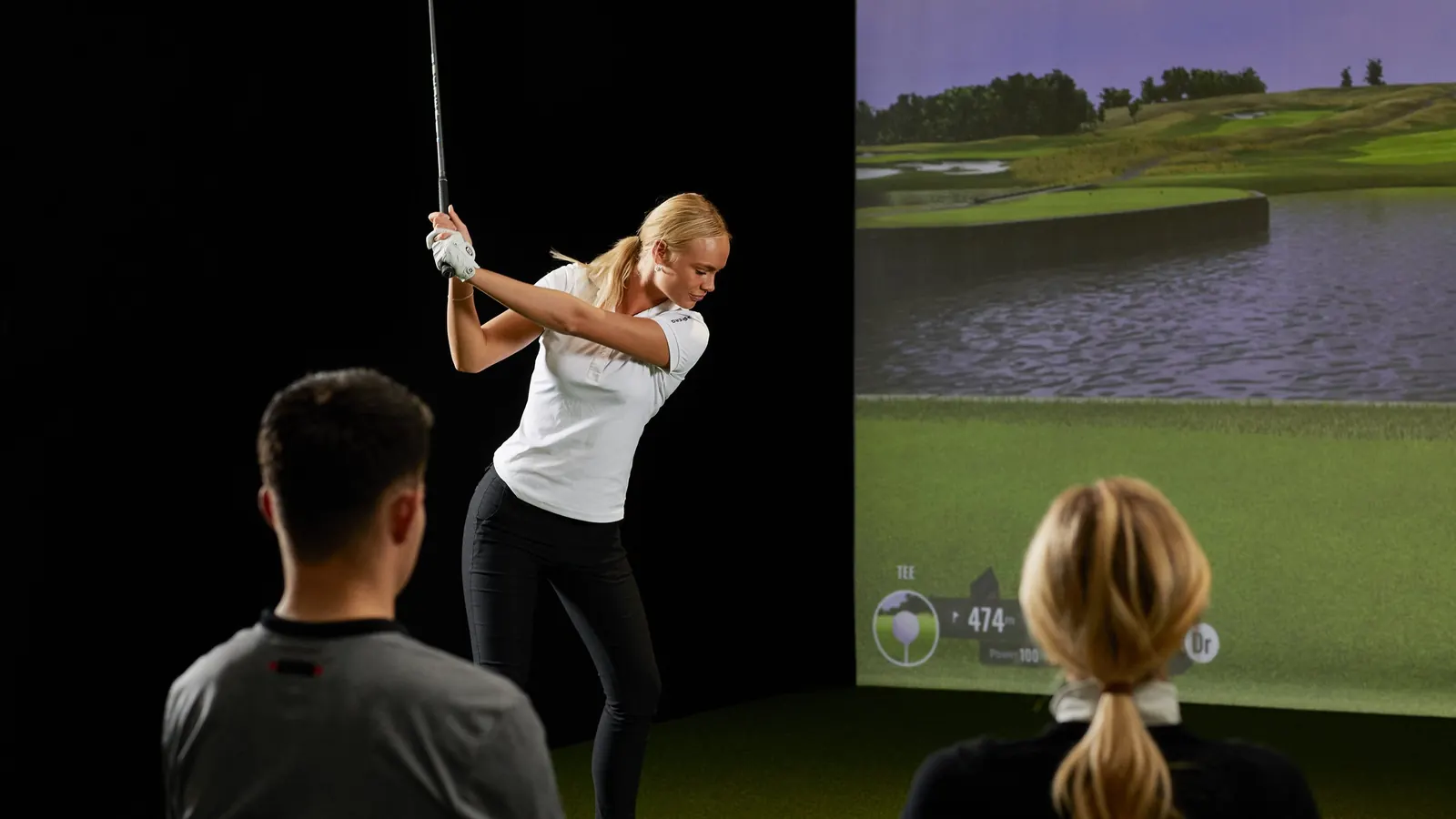 Golferin beim Schwung am Simulator, Trainer und Zuschauerin schauen zu
