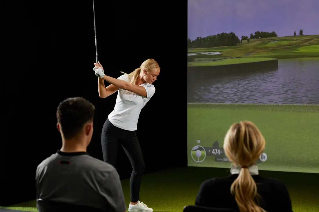 Golferin beim Schwung am Simulator, Trainer und Zuschauerin schauen zu